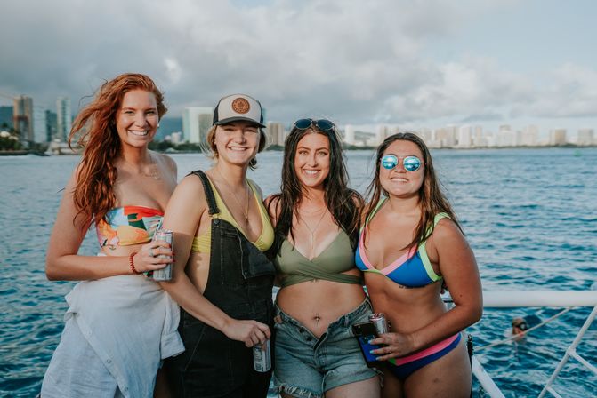 Four smiling women in colorful swimsuits and summer outfits posing on a boat with blue ocean and a coastal city skyline in the background, holding canned drinks and enjoying a sunny day