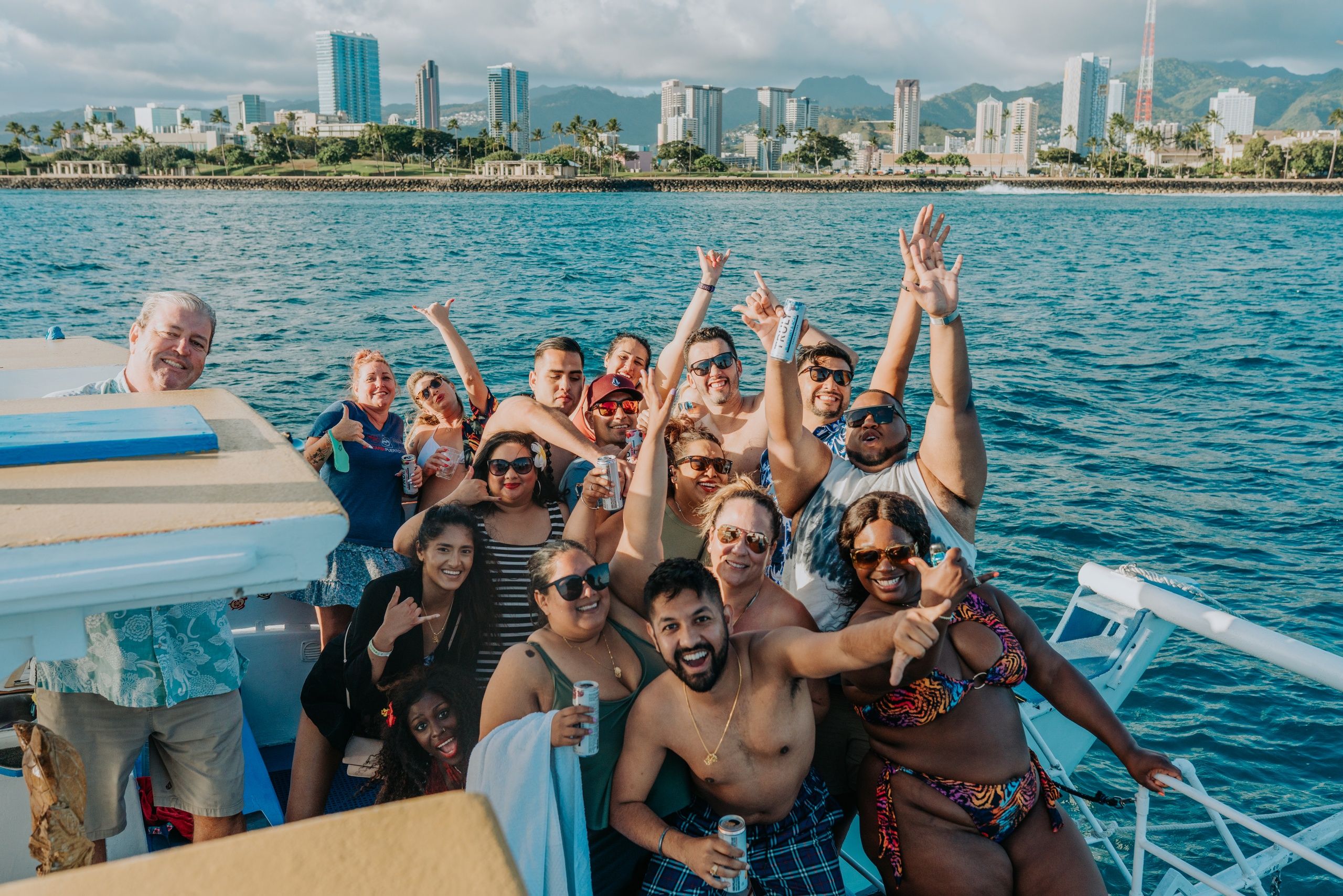 Group of friends enjoying a sunny boat party on the blue ocean, holding drinks with a tropical city skyline and mountains in the background.