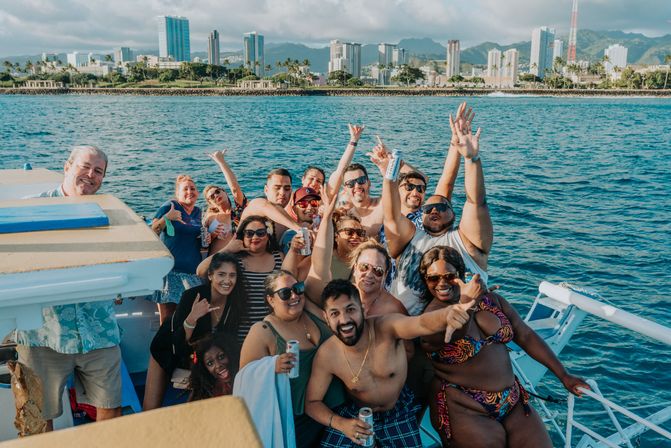 Group of friends enjoying a sunny boat party on the blue ocean, holding drinks with a tropical city skyline and mountains in the background.