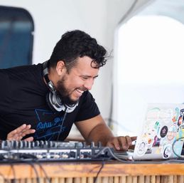 Smiling DJ with headphones leaning over a sticker-covered laptop and audio mixer on a sunlit wooden booth.