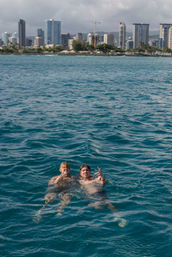 Two friends floating and waving in deep blue ocean off a palm-lined urban waterfront with high-rise city skyline in the background.