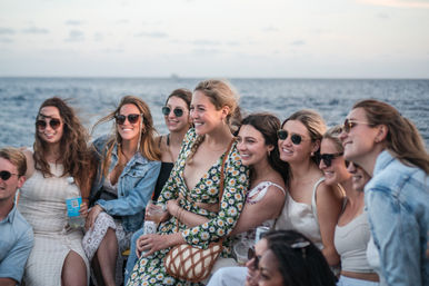 Cheerful group of friends on a boat at the ocean, smiling and posing together at sunset in summer dresses and sunglasses while holding drinks on a coastal outing.