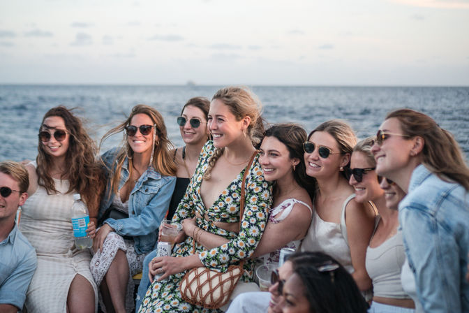 Cheerful group of friends on a boat at the ocean, smiling and posing together at sunset in summer dresses and sunglasses while holding drinks on a coastal outing.