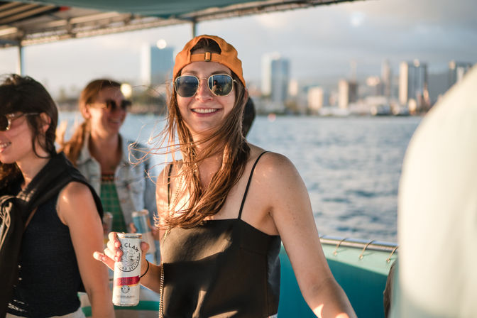Smiling woman in sunglasses and backward cap holding a canned drink on a sunny boat cruise with city skyline and water in the background