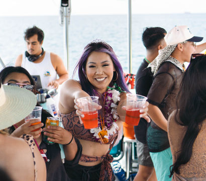 Smiling woman with purple hair, tiara and lei handing out red drinks at a crowded tropical boat party on the ocean