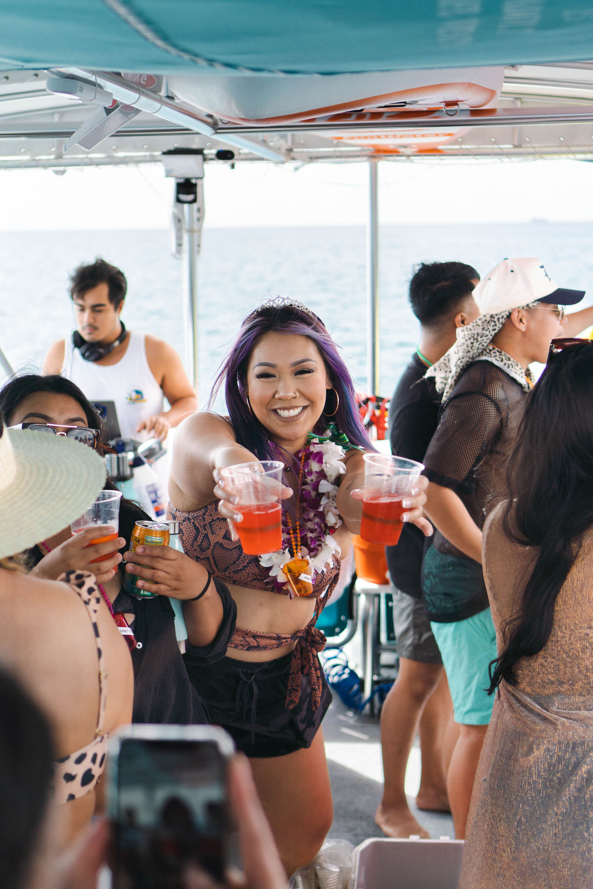 Smiling woman with purple hair, tiara and lei handing out red drinks at a crowded tropical boat party on the ocean