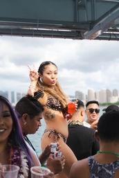 Smiling woman in a leopard-print bikini flashes a peace sign and holds a red drink aboard a crowded boat party, friends nearby and a coastal city skyline across the water.