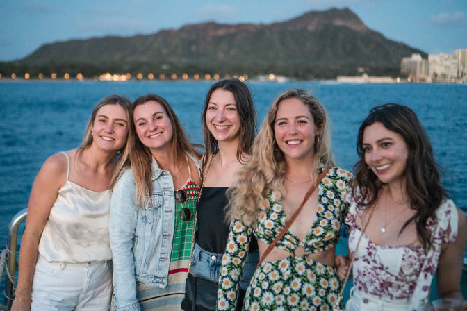 Five friends smiling on a boat at dusk with the blue ocean and Diamond Head crater and Waikiki skyline in the background, Honolulu, Hawaii.
