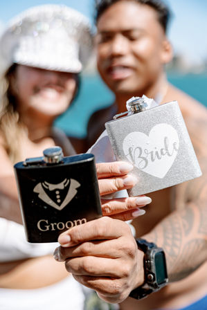Silver glitter 'Bride' flask and black 'Groom' flask held up in foreground while a smiling couple celebrates on a sunny turquoise beach