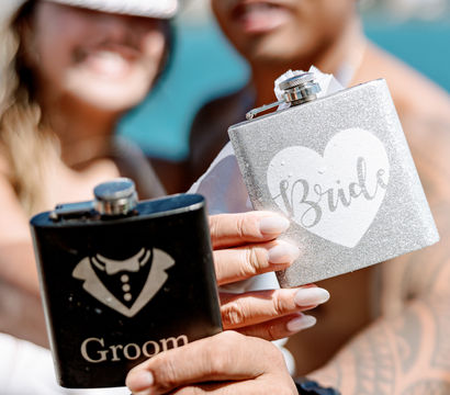 Silver glitter 'Bride' flask and black 'Groom' flask held up in foreground while a smiling couple celebrates on a sunny turquoise beach