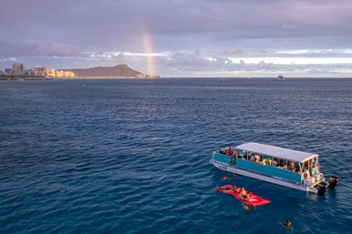 Passenger boat and swimmers on a red float in the blue Pacific off Waikiki, with Diamond Head, shoreline skyline and a rainbow at sunset in Hawaii