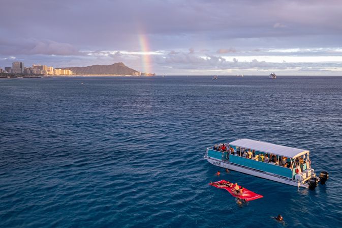 Passenger boat and swimmers on a red float in the blue Pacific off Waikiki, with Diamond Head, shoreline skyline and a rainbow at sunset in Hawaii