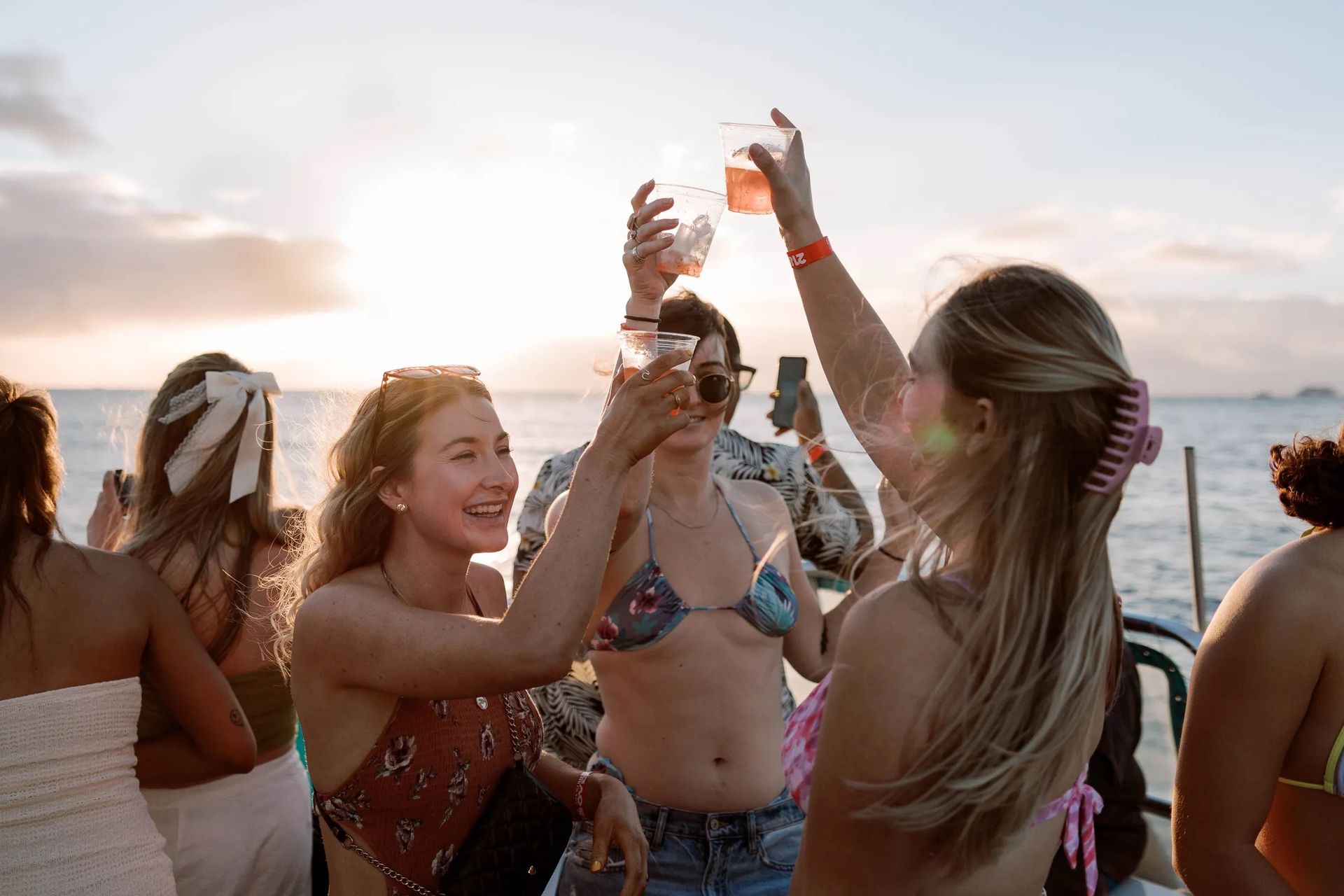 Group of friends toasting with plastic cups on a sunset boat cruise over the ocean, smiling women in swimsuits enjoying a coastal sunset party