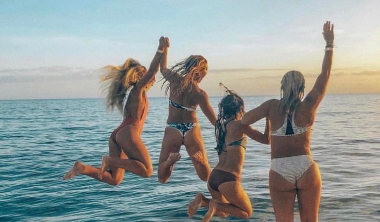 Four friends in bikinis jumping hand-in-hand into the ocean at sunset, playful beach vacation scene with calm blue water and a warm golden sky