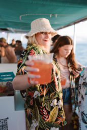 Person in a tropical print shirt and white bucket hat holding an orange cocktail toward the camera on a lively sunset boat party with the ocean in the background.