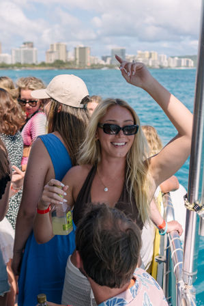 Smiling blonde woman in sunglasses holding a bottled drink and raising her arm aboard a crowded boat with turquoise ocean and a coastal city skyline in the background