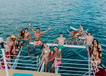 Young adults in swimwear partying on a boat and in clear blue ocean, some perched on outriggers and a paddleboard, smiling, waving and holding drinks on a sunny summer boat trip.