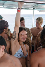 Smiling young woman in a light blue bikini raising her arm to grip a boat's overhead rail, revealing an upside-down portrait tattoo on her inner arm while surrounded by friends on a sunny ocean boat party.
