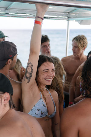 Smiling young woman in a light blue bikini raising her arm to grip a boat's overhead rail, revealing an upside-down portrait tattoo on her inner arm while surrounded by friends on a sunny ocean boat party.