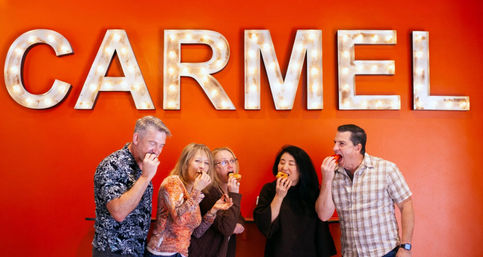 Five adults laughing and biting into pastries beneath oversized illuminated marquee letters on a bright orange wall.
