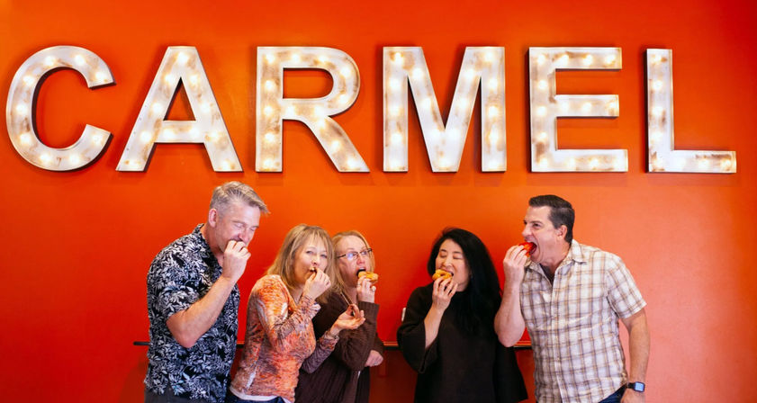 Five adults laughing and biting into pastries beneath oversized illuminated marquee letters on a bright orange wall.
