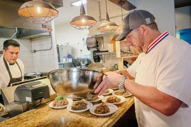Chef in white jacket and cap plating multiple pasta dishes from a large stainless-steel bowl on a restaurant pass, with a line cook, pendant lights and tiled kitchen in the background.