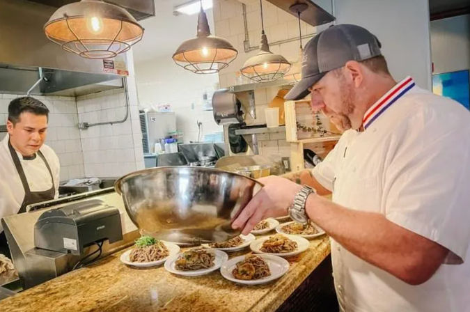 Chef in white jacket and cap plating multiple pasta dishes from a large stainless-steel bowl on a restaurant pass, with a line cook, pendant lights and tiled kitchen in the background.