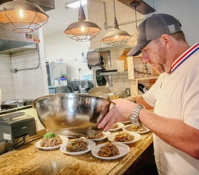 Chef in white jacket and cap plating multiple pasta dishes from a large stainless-steel bowl on a restaurant pass, with a line cook, pendant lights and tiled kitchen in the background.