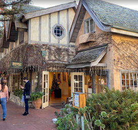 Quaint Tudor-style storefront with ivy-covered stucco, open double doors revealing a Christmas tree, icicle string lights and pedestrians on a brick walkway.