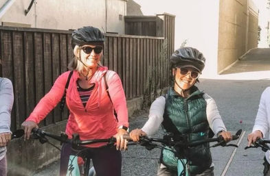 Two smiling cyclists in helmets and sunglasses on a sunny residential street — one in a pink jacket and the other in a green vest — holding bike handlebars by a wooden fence and alley.