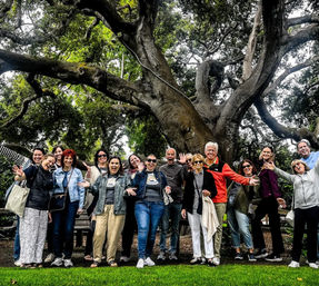 Cheerful group of adults posing playfully beneath a massive sprawling oak tree in a park, with lush green grass in the foreground and twisting branches overhead.