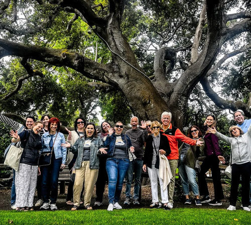 Cheerful group of adults posing playfully beneath a massive sprawling oak tree in a park, with lush green grass in the foreground and twisting branches overhead.