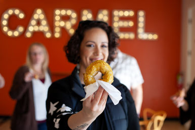 Smiling person holds a shiny glazed donut on a napkin toward the camera inside a bright orange donut shop with marquee lights and friends blurred in the background.