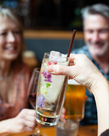Cheers-worthy craft cocktail with a tall glass, floral ice cube and brown straw raised for a toast at a lively bar, blurred smiling friends and beer in the background