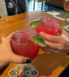 Two bright pink cocktails with crushed ice and lime wedges clinking over a wooden bar table, hands holding glasses and a flamingo-patterned coaster visible in an indoor bar setting.