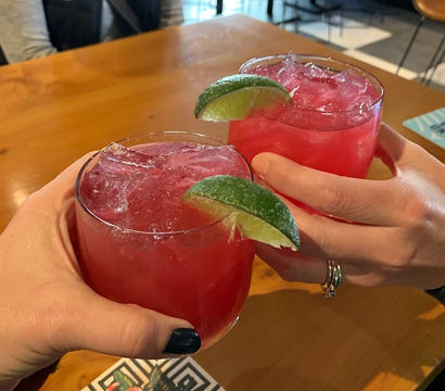 Two bright pink cocktails with crushed ice and lime wedges clinking over a wooden bar table, hands holding glasses and a flamingo-patterned coaster visible in an indoor bar setting.