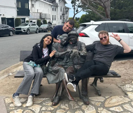 Three friends laugh and pose with a life-size bronze bench sculpture of an elderly couple on a residential street, with parked cars and houses in the background.