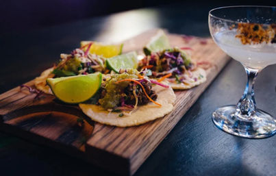 Three street-style tacos with shredded slaw, green salsa and lime wedges on a wooden board beside a cocktail glass on a dark bar counter