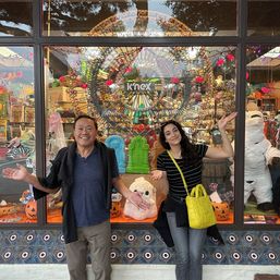 Two smiling people with arms outstretched pose in front of a colorful storefront Halloween window display featuring a large Ferris-wheel-style model, jack-o’-lanterns, tombstones, a wrapped mummy figure, patterned tile base, and a bright yellow shoulder bag.