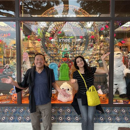Two smiling people with arms outstretched pose in front of a colorful storefront Halloween window display featuring a large Ferris-wheel-style model, jack-o’-lanterns, tombstones, a wrapped mummy figure, patterned tile base, and a bright yellow shoulder bag.