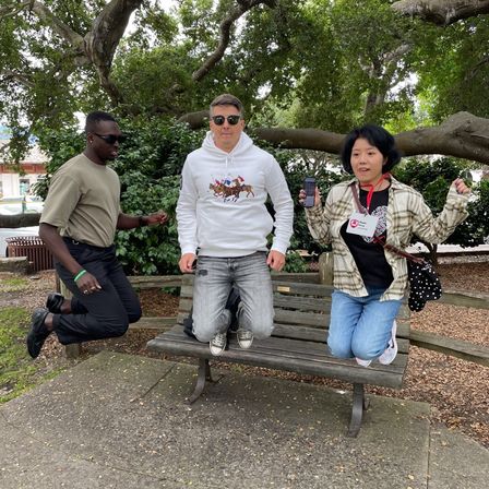 Three people mid-jump on a wooden park bench in a city park beneath a sprawling tree, center person in a white hoodie with horse graphic, left in a khaki tee and sunglasses, right holding a phone and wearing a lanyard — lively daytime outdoor scene.