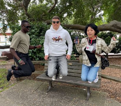Three people mid-jump on a wooden park bench in a city park beneath a sprawling tree, center person in a white hoodie with horse graphic, left in a khaki tee and sunglasses, right holding a phone and wearing a lanyard — lively daytime outdoor scene.