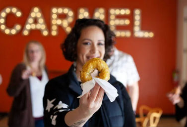 Smiling person offering a glossy glazed donut on a napkin inside a bright red local donut shop with marquee lights and blurred customers in the background.