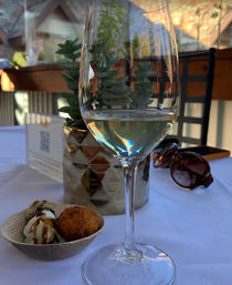Stemmed glass of white wine on a white-tablecloth al fresco patio table with sunset reflections, a potted succulent centerpiece, small appetizer bowl with a fried croquette and mozzarella drizzled in balsamic, and sunglasses nearby