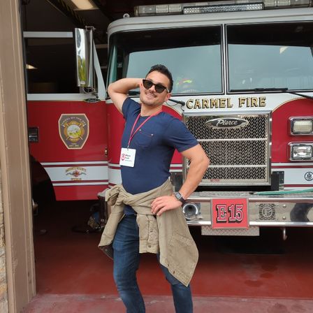 Person in sunglasses striking a playful pose in front of a red fire engine inside a fire station, wearing a navy T‑shirt, jacket tied at the waist and a visitor lanyard.