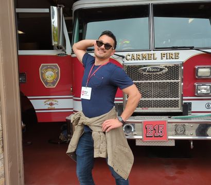 Person in sunglasses striking a playful pose in front of a red fire engine inside a fire station, wearing a navy T‑shirt, jacket tied at the waist and a visitor lanyard.