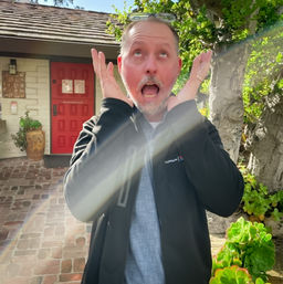 Playful person with hands to their cheeks and a surprised expression on a sunlit brick patio in front of a red door, potted plants, succulents and a tree with a visible lens flare.