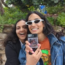 Two friends laughing in a garden selfie by a stone wall and succulents; one wearing white cat‑eye sunglasses and a denim jacket holds a phone showing a childhood photo.