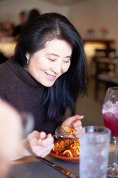 Smiling woman with long dark hair enjoying saucy pasta at a cozy café table, fork in hand and a pink drink nearby