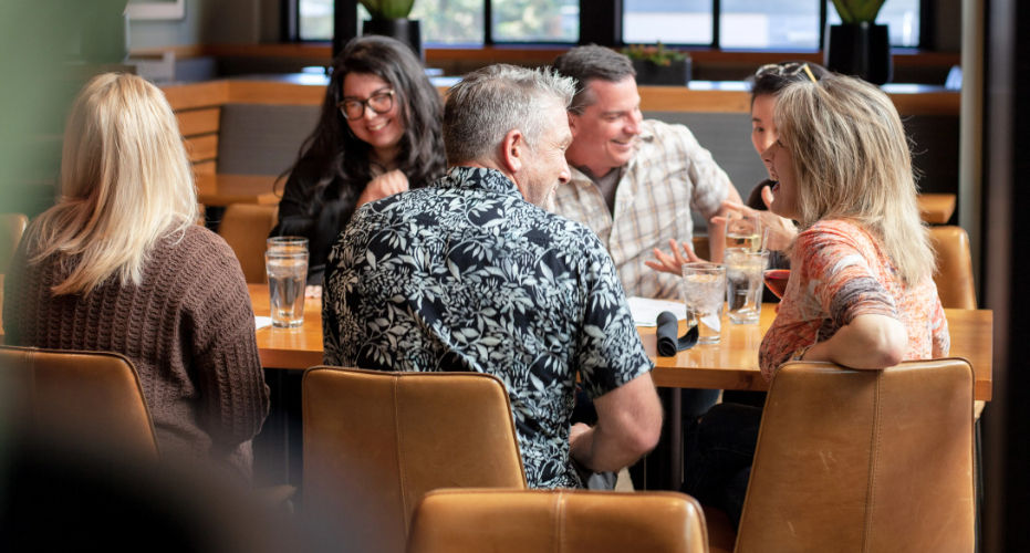 Group of friends laughing and chatting over drinks at a cozy indoor restaurant table in a casual dining setting.